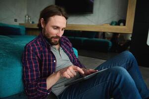Relaxed smiling Caucasian man in casual denim and checkered shirt reads news on digital tablet, surfs internet, sitting on floor at home leaning back on a sofa. Leisure time, recreation, lifestyle photo