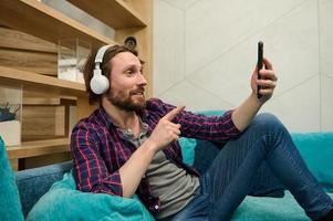 Handsome relaxed bearded Caucasian man wearing wireless headphones, talking by video call, looking at web camera on the smartphone in his hand, relaxing lying down on a sofa at home photo