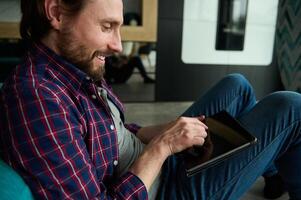 Close-up of a handsome Caucasian bearded man in casual clothes works at home on a digital tablet. Digital electronic gadgets in everyday life. Internet, online, remote work, connection concept photo