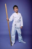 Handsome confident child boy in white kimono with wooden weapon bokken over purple background with copy space. Oriental martial arts concept, Aikido photo