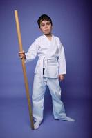 hermoso confidente niño chico en blanco kimono con de madera arma bokken terminado púrpura antecedentes con Copiar espacio. oriental marcial letras concepto, aikido foto