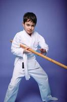 un Adolescente chico en un blanco kimono peleas con un de madera espada en aikido formación en un púrpura antecedentes. sano estilo de vida y marcial letras concepto foto