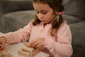 Close-up portrait of a charming cute adorable little preschool girl in pink sweatshirt playing with wooden blocks, building structures. Fine motor skills development and educational leisure concept photo