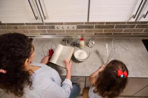 Overhead view of mother and daughter cooking food together while standing at the kitchen countertop. Mom holding recipe book ready to teach her adorable baby girl how to cook dough photo