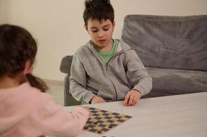 linda pequeño niña disfrutando intelectual tablero juego con su más viejo hermano, colocación juego de damas en el tablero de ajedrez. lógica desarrollo, inteligente ocio, educación, entretenimiento y inteligente pasatiempo concepto foto