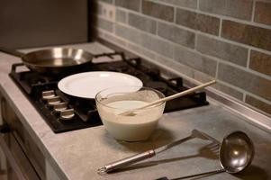 Wooden spoon in glass bowl with pancake batter, stainless steel kitchen utensils on kitchen countertop, white plate and frying pan on a black stove photo