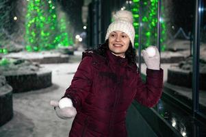 contento joven mujer vistiendo calentar ropa, blanco lana sombrero y mitones, jugando en bola de nieve peleas en un Nevado invierno noche foto