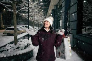 encantador mujer con compras bolsas, capturas copos de nieve mientras camina a lo largo un Nevado calle iluminado por fiesta guirnaldas foto