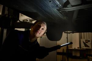 Technician checking modern car at automobile repair shop. Auto mechanic using a flashlight lamp for inspecting the underbody of the lifted car in a car service. Automobile maintenance concept photo