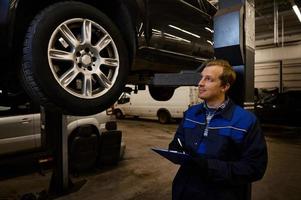 auto mecánico, técnico en uniforme haciendo el Lista de Verificación en portapapeles para reparando el coche levantado en un izar en el reparar tienda durante garantía coche mantenimiento y Servicio foto