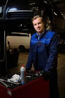 Professional portrait of a handsome Caucasian man, auto mechanic, technician, car engineer standing near a box with set of tools for repairing a lifted automobile on a hoist in the repair shop garage photo