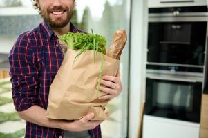 Close-up eco paper bag, craft cardboard packet with greens, salads, healthy food, whole grain baguette bread in hands of handsome caucasian guy smiling beautiful toothy smile standing in home kitchen photo