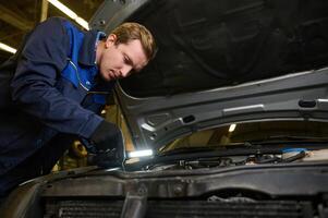 Technician, garage mechanic using flashlight luminous lamp while inspecting a car under hood in repair shop. Auto service, car maintenance concept photo