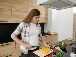 sereno embarazada mujer en blanco camiseta y a cuadros camisa preparando comida en cocina isla desde Fresco crudo vegano ingredientes, pone rebanadas campana pimienta dentro un bol. sano comiendo para sano maternidad foto