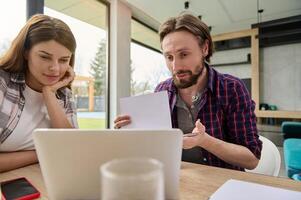 Young couple talking by video call, sitting in front of a laptop, and showing documents to the the web camera. Online conference, remote work from home, internet, freelance occupation concept photo