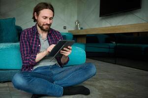 Confident handsome bearded European man in casual jeans and plaid shirt holding digital tablet and looking at camera, sitting on floor with back against sofa while working from home photo