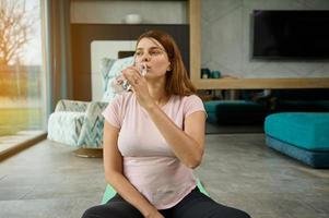 Young woman in anticipation of a baby sits on a yoga mat in lotus pose, drinks water while relaxing after a light workout at home. Pregnancy and sport concept. Pregnant active woman exercising at home photo