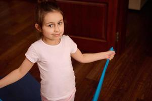 Lovely school girl in sportswear, standing with elastic fitness band while working out indoors, smiles looking at camera photo