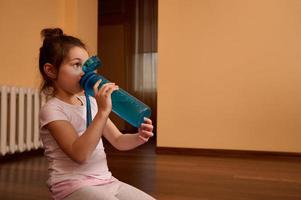 adorable niño niña en blanco camiseta, sentado en aptitud estera y Bebiendo agua desde azul Deportes botella foto