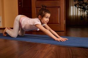 Side portrait of active sporty child girl stretching body and back on fitness mat, practicing yoga, exercising indoors photo