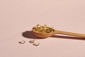 Still life with wooden spoon full of soft gel capsules with golden color oil, on isolated pink background photo