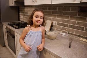 retrato de un contento adorable hermosa pequeño caucásico niña en azul vestir sonriente lindo mirando a cámara, propensión su brazo en un cocina encimera mientras preparando masa a hogar cocina foto