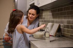 adorable contento madre sonrisas a su hija, linda pequeño niña mezcla el ingredientes en un cuenco mientras preparando tortita masa. maternidad, mamá enseñando su hija a cocinar panqueques para carnaval martes foto