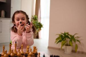 Adorable little girl holding chess pieces and cutely smiles with cheerful toothy smile looking at camera while playing chess with her brother in the home interior. Chess game for clever mind photo