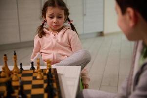 bonito 4 4 años antiguo pequeño niña con trastornado Mira sentado siguiente a su hermano y aprendizaje ajedrez juego foto