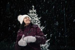 un encantador mujer en calentar ropa, disfrutando Navidad atmósfera al aire libre en Nevado noche. nevada. alegre Navidad foto