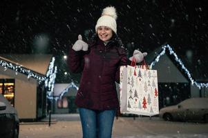 Charming woman experiencing happiness, showing thumbs up, smiling cheerfully to the camera, on the street at snowy night photo