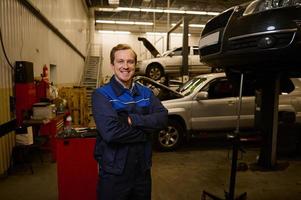 Confident professional portrait of a handsome auto mechanic in uniform smiling with a toothy smile, standing in a car service garage with a lifted car in the background photo