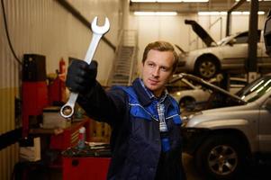 Professional portrait of a young Caucasian auto mechanic in uniform holding a wrench while standing at his workplace in a car service. Car repair and maintenance. photo