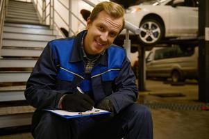 retrato de un joven sonriente caucásico auto mecánico sentado en escalera en el reparar tienda y mirando a cámara mientras escritura listas de verificación en portapapeles, inspeccionando el coche en taller. foto