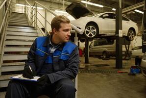 Caucasian mechanic sitting on stairs in the repair shop against cars with open hood, lifted on hoist, writing checklists on clipboard. Mechanical maintenance engineer working in automotive industry. photo