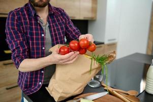 Close-up of a Caucasian man taking out vegetables and healthy food from a paper eco bag, holding a sprig of fresh tomatoes in his hands, putting it on the kitchen table. Healthy eating concept photo