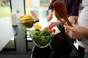 Focus on a glass bowl full of healthy greens, lettuce leaves and fresh vegetables on the background of a happy pregnant couple preparing healthy raw vegan salad on the kitchen island photo