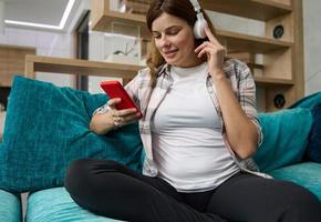 Nice adorable lovely cheerful positive optimistic pregnant woman, future mom in casual wear, sitting on the couch at home enjoying classical music and swiping websites and mobile apps on smartphone photo