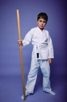 Learning aikido, Oriental martial arts concept. Cute boy in a white kimono with a wooden weapon in his hand isolated on purple background copy space photo