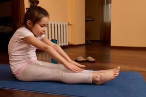 pequeño niña haciendo extensión ejercicios brazos y piernas en yoga estera, practicando gimnasia y aptitud actividad a hogar. foto