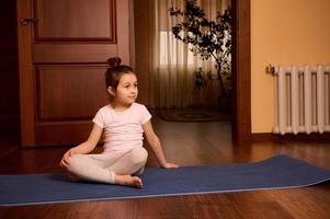 Adorable Caucasian 5 years old little child girl in active wear, sitting on yoga mat at cozy home interior. photo
