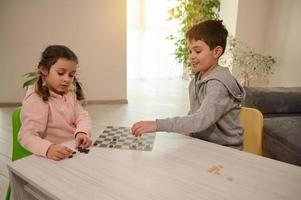 Two adorable Caucasian elementary aged kids, boy and girl, brother and sister having great time playing checkers board game together at home interior photo