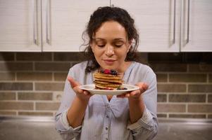 Beautiful woman holding a plate of delicious homemade freshly baked pancakes garnished with wild berries. Charming young dark-haired curly housewife with cooked pancakes for Shrovetide. Shrove Tuesday photo