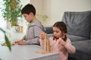 Creative European little girl sitting next to her brother and focused on construction of complex tall structure from wooden blocks, . Fine motor skills, concentration and educational leisure concept photo