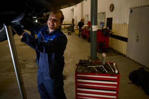 hermoso caucásico joven hombre, coche ingeniero, técnico, auto mecánico reparando un levantado moderno automóvil en un izar en el reparar tienda cochera. automóvil mantenimiento y auto Servicio concepto foto