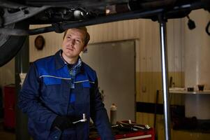 auto mecánico en uniforme inspeccionando un levantado coche en un izar, mientras haciendo Lista de Verificación para garantía reparando automóvil en el taller garaje foto