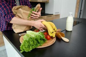 Overhead view of an unrecognizable man unpacking eco cardboard bag with healthy raw vegan food and a whole grain baguette, putting it on a kitchen table. Healthy food and lifestyles concept photo