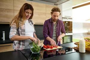 hermosa embarazada mujer poniendo Rúcula y ensalada hojas dentro un vaso cuenco mientras su amoroso marido rebanar Tomates en un corte tablero. contento Pareja Cocinando juntos a hogar cocina. maternidad foto