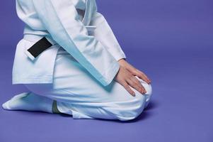 Cropped view of child aikido wrestler in white kimono sitting in stance with hands on knees isolated on purple background with copy space. Concept of Oriental Martial Arts practice photo