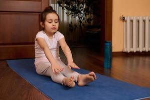 determinado pequeño niña 5-6 años viejo, hacer ejercicio descalzo en yoga estera, extensión su cuerpo, haciendo gimnástico ejercicios foto